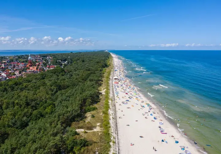 Breiter Sandstrand mit vielen Badegästen, gesäumt von einem dichten Kiefernwald und einer kleinen Stadt. Ein echter Geheimtipp an der polnischen Ostsee.