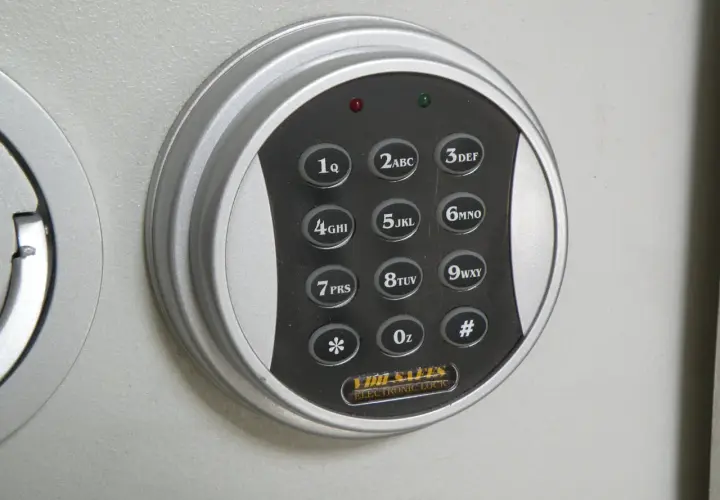 Elektroniczna klawiatura szyfrowa sejfu z przyciskami cyfrowymi i symbolami. W tle widoczne są metalowe tuleje mocujące.