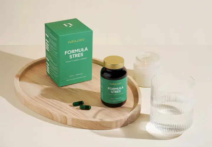 Zielone opakowanie i butelka suplementu diety "Formuła Stres", dwa kapsułki, szklanka wody i świeca.