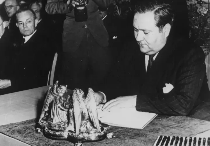 Carlo Schmid sitzt an einem Tisch und blickt auf ein Buch. Ein Fotograf hält eine Kamera.