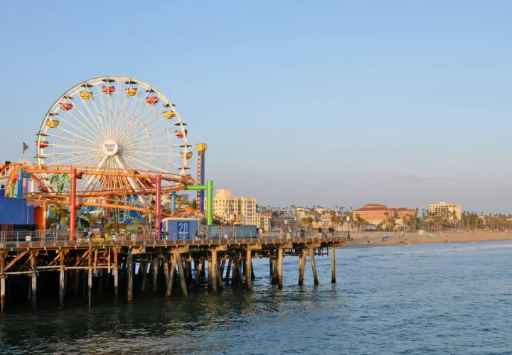 Der Santa Monica Pier mit seinem Riesenrad und Achterbahn ragt ins Meer. Im Hintergrund erstreckt sich der sonnige los angeles strand mit Palmen und Gebäuden.