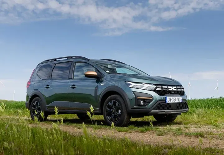Un'auto familiare verde scuro, con barre sul tetto e dettagli color rame, parcheggiata su un sentiero sterrato in un campo con mulini a vento sullo sfondo.