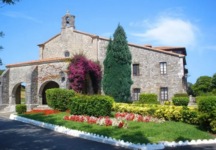 El **santuario de la Barquera**, con su fachada de piedra y arcos, se adorna con vibrantes buganvillas y un jardín florido bajo un cielo azul.