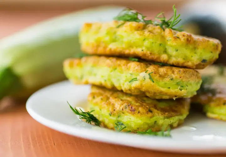 Frittelle di zucchine dorate e soffici, guarnite con aneto fresco, impilate su un piatto bianco. Sullo sfondo una zucchina intera.