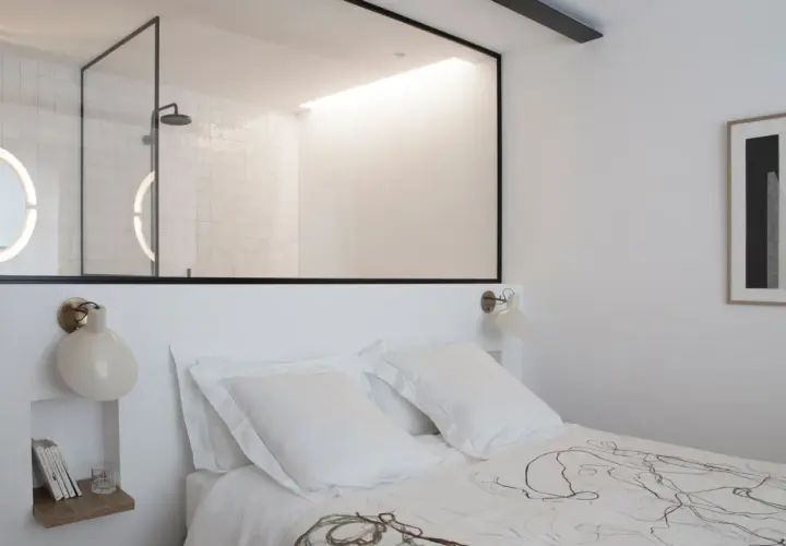 Stanza buia con letto bianco, una finestra tra due stanze rivela un bagno moderno con doccia e specchi luminosi.