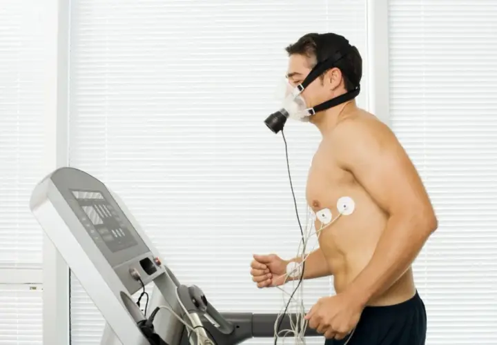 Mężczyzna w masce biegający na bieżni, podłączony do aparatury EKG. Wyniki testu VO2max w tabeli na ekranie bieżni.