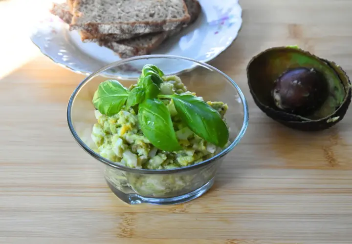 Pyszna pasta jajeczna z awokado, udekorowana bazylią, obok połówka awokado i kromki chleba.