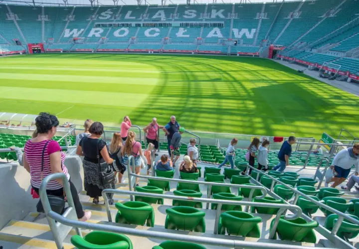 Grupa ludzi wchodzi na zielone trybuny stadionu. W tle widać murawę i napis "WROCŁAW".