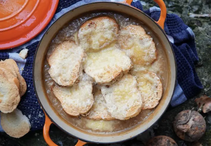 Zuppa di cipolle fumante in una casseruola arancione, con crostini dorati e formaggio fuso. Accanto, pane tostato e cipolle crude.