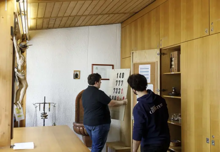Zwei Männer im katholischen Priesterseminar hängen eine Liste mit Zahlen auf. Ein Kruzifix hängt an der Wand.