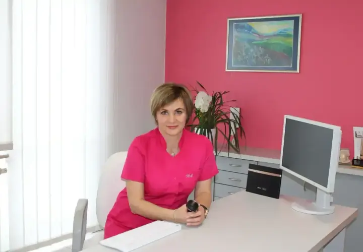 Lekarka w różowym uniformie siedzi przy biurku w gabinecie estetycznym.