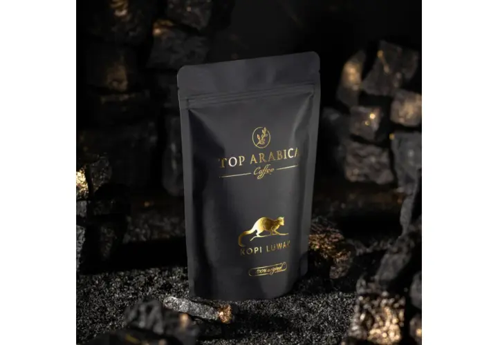 Czarna torebka z napisem "TOP ARABICA Coffee Kopi Luwak" i złotym rysunkiem zwierzęcia. To najdroższa kawa rozpuszczalna, otoczona węglem i złotymi drobinkami.