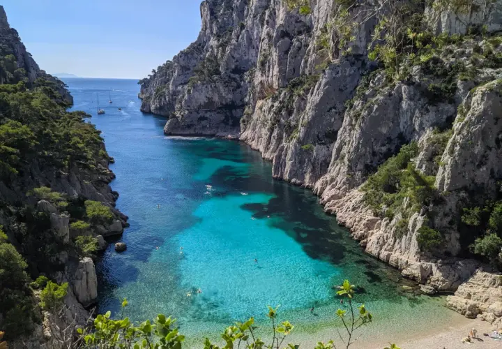 Aguas turquesas y cristalinas en el **calanque d'en Vau**, rodeado de imponentes acantilados rocosos y vegetación. Gente nadando y barcos a lo lejos.
