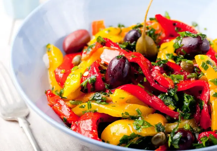 Insalata colorata di peperoni in agrodolce, con olive nere, capperi e prezzemolo fresco, servita in una ciotola blu.