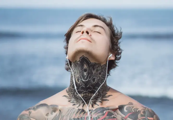 Mężczyzna z tatuażem na obojczyku i szyi, słuchający muzyki na plaży.