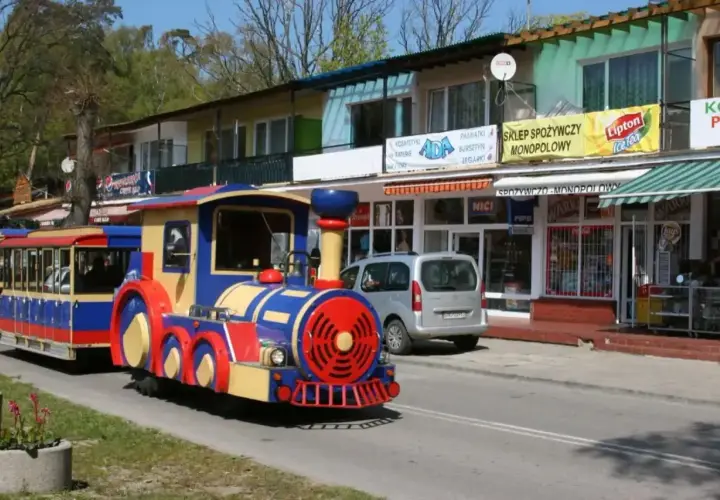 Kolorowa kolejka turystyczna jedzie ulicą. W tle sklepy, jeden z nich reklamuje "autobus 870 Gdańsk cennik".