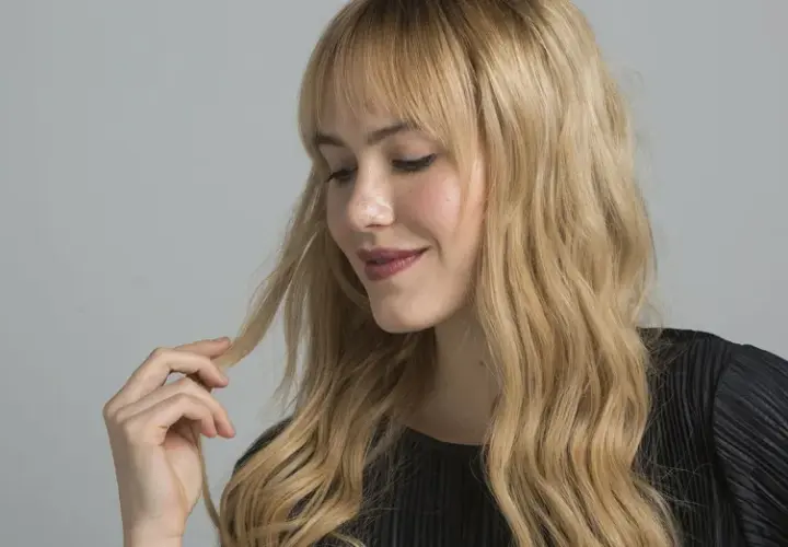 Mujer con cabello rubio ondulado y flequillo, tocando un mechón. El matizar el pelo realza su brillo y tono natural.