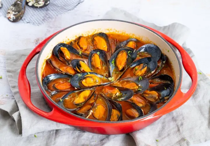 Mejillones en salsa roja, servidos en una cazuela roja, con perejil picado espolvoreado.