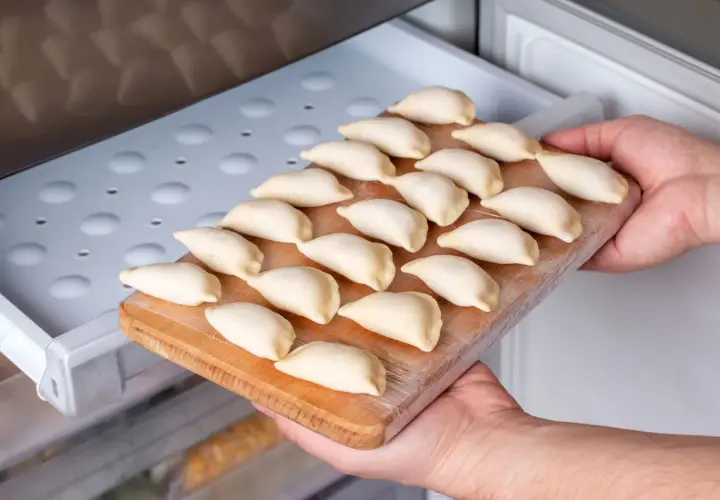 Ręce wkładają deskę z surowymi pierogami do zamrażarki. To prosty sposób, jak przechować ciasto na pierogi na później.