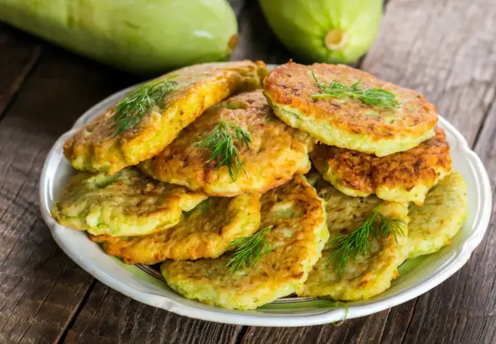 Pile de beignets de courgette dorés et moelleux, garnis de brins d'aneth frais, sur une assiette blanche. Deux courgettes vertes floues en arrière-plan.