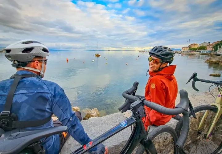 Two cyclists enjoy a break during bike guided tours in the Mediterranean, with boats on the calm sea and a coastal town in the background.