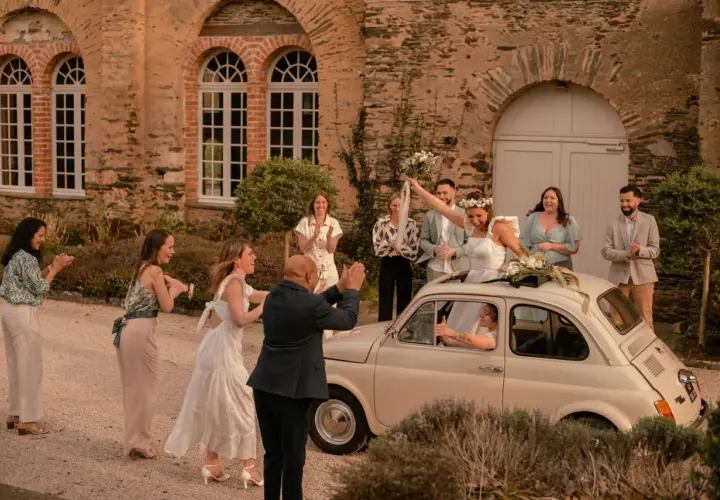 Mariage Dolce Vita : la mariée, radieuse, lève son bouquet depuis une Fiat 500. Ses invités applaudissent devant un manoir en pierre.