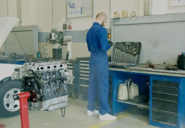Mechanik w niebieskim kombinezonie przy silniku samochodu. Obok skrzynka z narzędziami, płyny i kanistry. Unikaj mandatu za brak przeglądu, dbaj o auto.