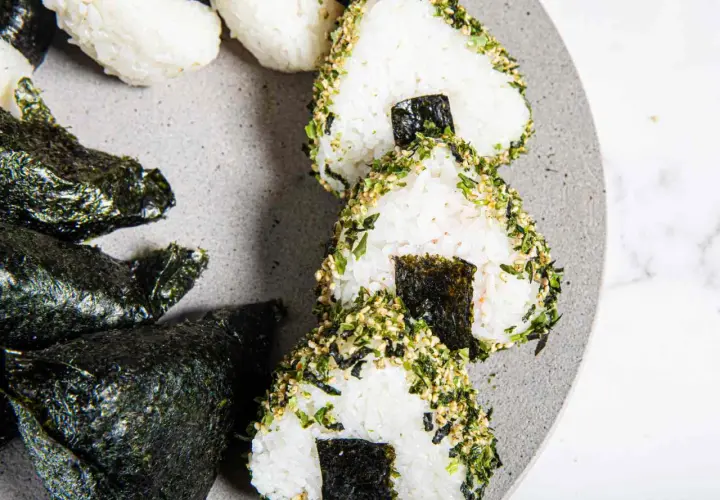 Triangular sushi rice balls, some wrapped in nori, others coated in sesame seeds and herbs, are artfully arranged on a grey plate.