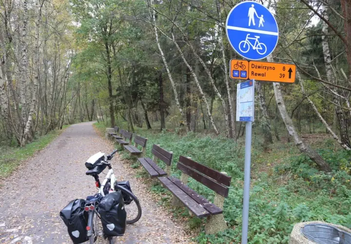 Ścieżka rowerowa Velo Baltica w lesie, z ławkami i znakiem wskazującym Dźwirzyno i Rewal. Długość trasy zachęca do podróży.