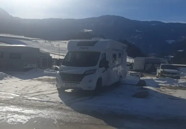 Kampery i przyczepy kempingowe na śniegu, gotowe na całoroczny camping w górach. Słońce przebija się przez chmury, oświetlając zimowy krajobraz.