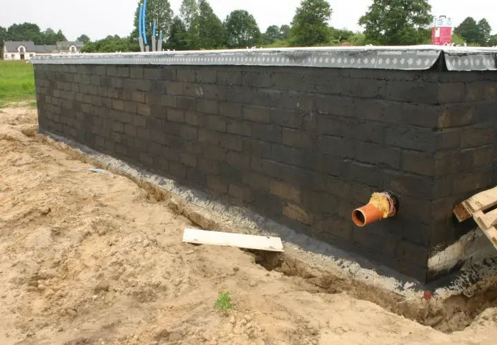 Pomarańczowa rura kanalizacyjna przechodzi przez fundament z czarnej cegły, przygotowywany do dalszych prac budowlanych.