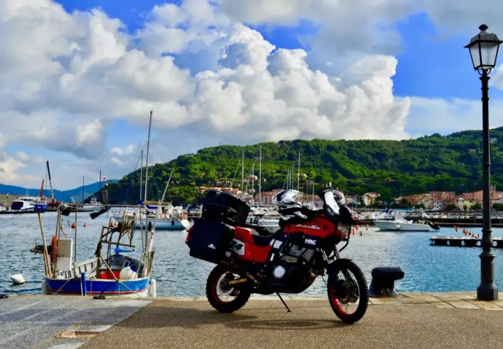 Moto rossa pronta a partire dal porto azzurro dell'Elba, con barche a vela e colline verdi sullo sfondo.
