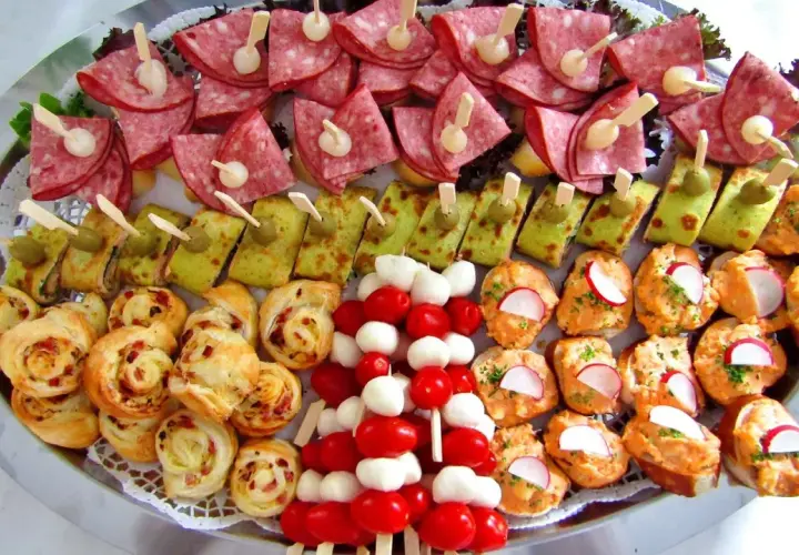 Talerz pełen pysznych przystawek na zimno: koreczki z pomidorków i mozzarelli, roladki z tortilli, kanapeczki z pastą i rzodkiewką, a także zawijane ciasteczka.
