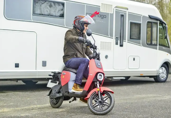 Un homme casqué conduit un scooter rouge devant un camping-car. Ce scooter est parfait pour explorer les environs lors de vos voyages en camping-car.