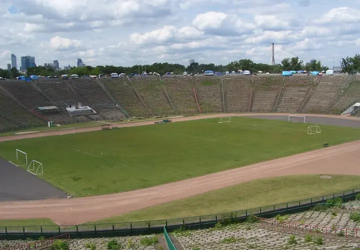 Stadion z PRL z murawą, bramkami i trybunami, w tle panorama Warszawy.