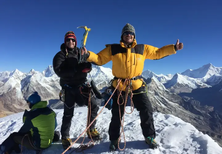 Dwóch alpinistów na szczycie Mera Peak, jeden z czekanem, drugi z kciukiem w górę. W tle ośnieżone góry.