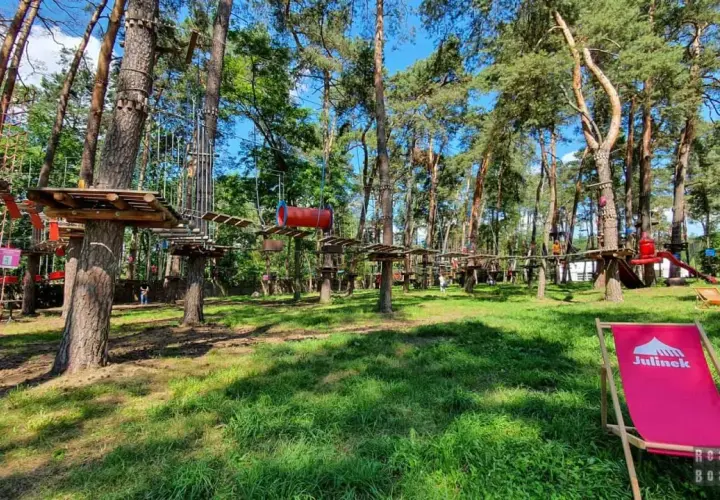 Park linowy w lesie z drewnianymi platformami i przeszkodami na drzewach. Na pierwszym planie różowy leżak.