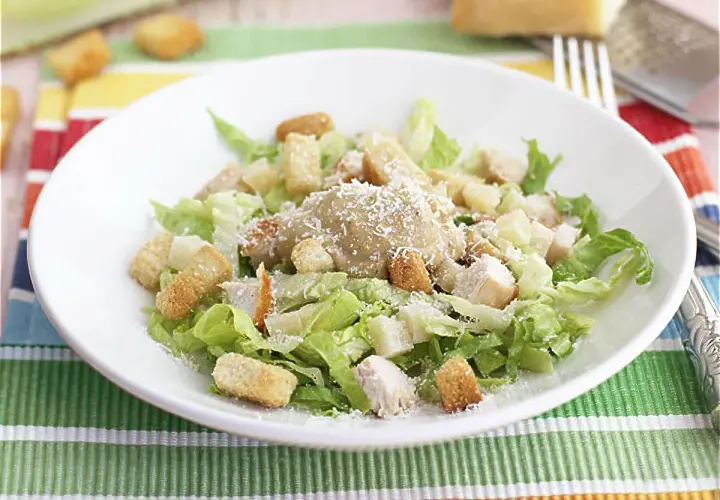 Una deliciosa ensalada cesar con pollo, crutones crujientes y queso parmesano rallado, servida en un plato blanco sobre un mantel colorido.