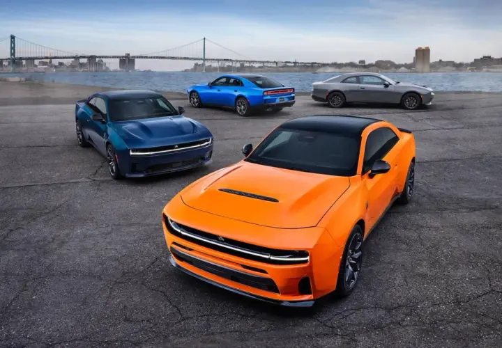 Pomarańczowy i niebieski Dodge Charger Hellcat oraz szary coupe stoją nad wodą z mostem w tle.