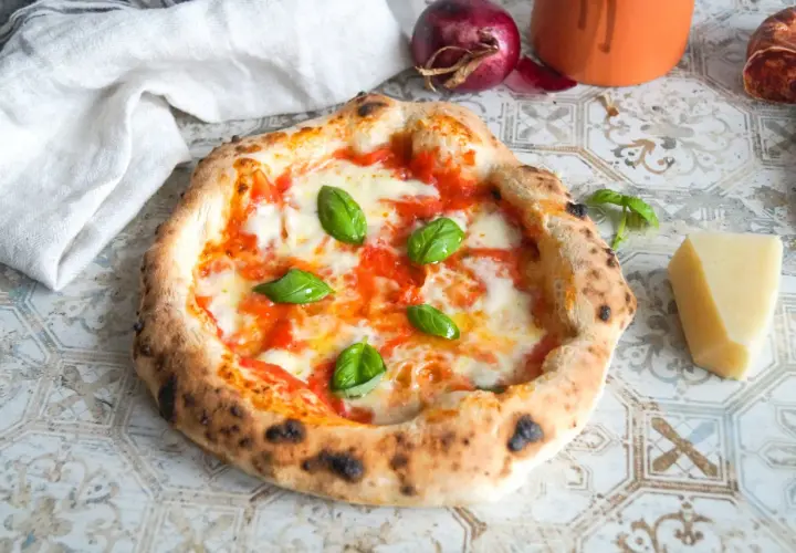 Eine authentische Pizza Napoletana mit frischem Basilikum, geschmolzenem Mozzarella und Tomatensauce. Daneben ein Stück Parmesan und eine rote Zwiebel.