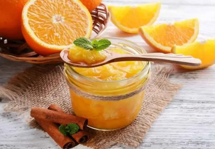 Tarro de mermelada de naranja casera, con naranjas frescas y canela en rama. Una cucharada muestra la textura.