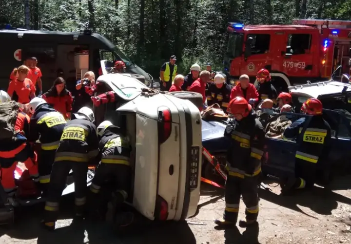 Akcja ratunkowa ksrg po wypadku. Strażacy wyciągają poszkodowanych z przewróconego samochodu. W tle wozy strażackie i karetka.