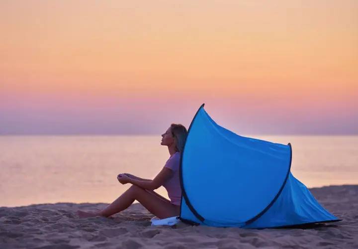 Kobieta siedzi na plaży przy rozłożonym namiocie plażowym. Zachód słońca nad morzem. Jak złożyć namiot plażowy instrukcja.