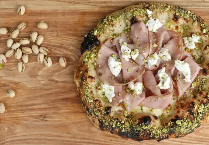 Pizza mit Mortadella, Pistazien und cremigem Käse, daneben einige Pistazien und eine Zitronenspalte.