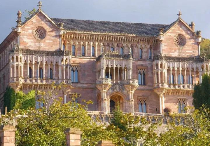 El **Palacio de Comillas**, una joya arquitectónica de piedra rosada, se alza majestuoso entre la vegetación, con sus balcones y rosetones detallados.