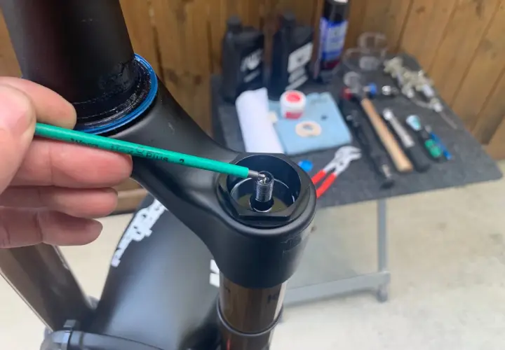 A hand uses a green hex wrench to adjust a bolt on a black bicycle fork, part of a Fox 34 service. Tools and lubricants are visible in the background.