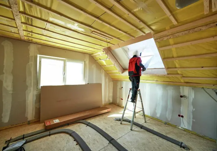 Lavoratore su scala installa isolamento giallo nel sottotetto, una scelta chiave per capire se meglio isolare il tetto o il sottotetto.