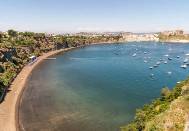 Vue d'une baie pittoresque, avec une plage de sable noir, des falaises verdoyantes et une multitude de bateaux dans l'eau bleue. Un paysage qui rappelle la voisine de Capri.