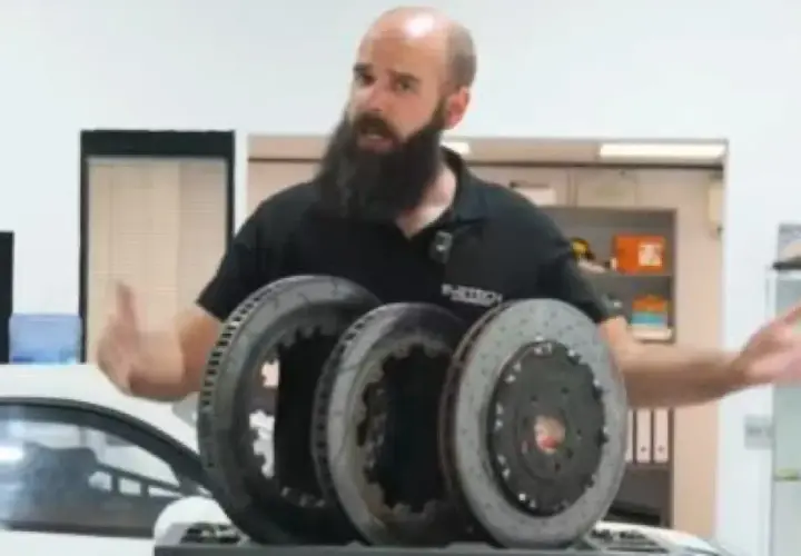 Hombre con barba muestra tres discos de freno, uno de ellos con perforaciones, para mejorar la refrigeración en un coche deportivo.