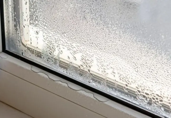 Gotas de agua cubren el cristal de la ventana, indicando condensación. El alféizar muestra pequeñas acumulaciones de agua.